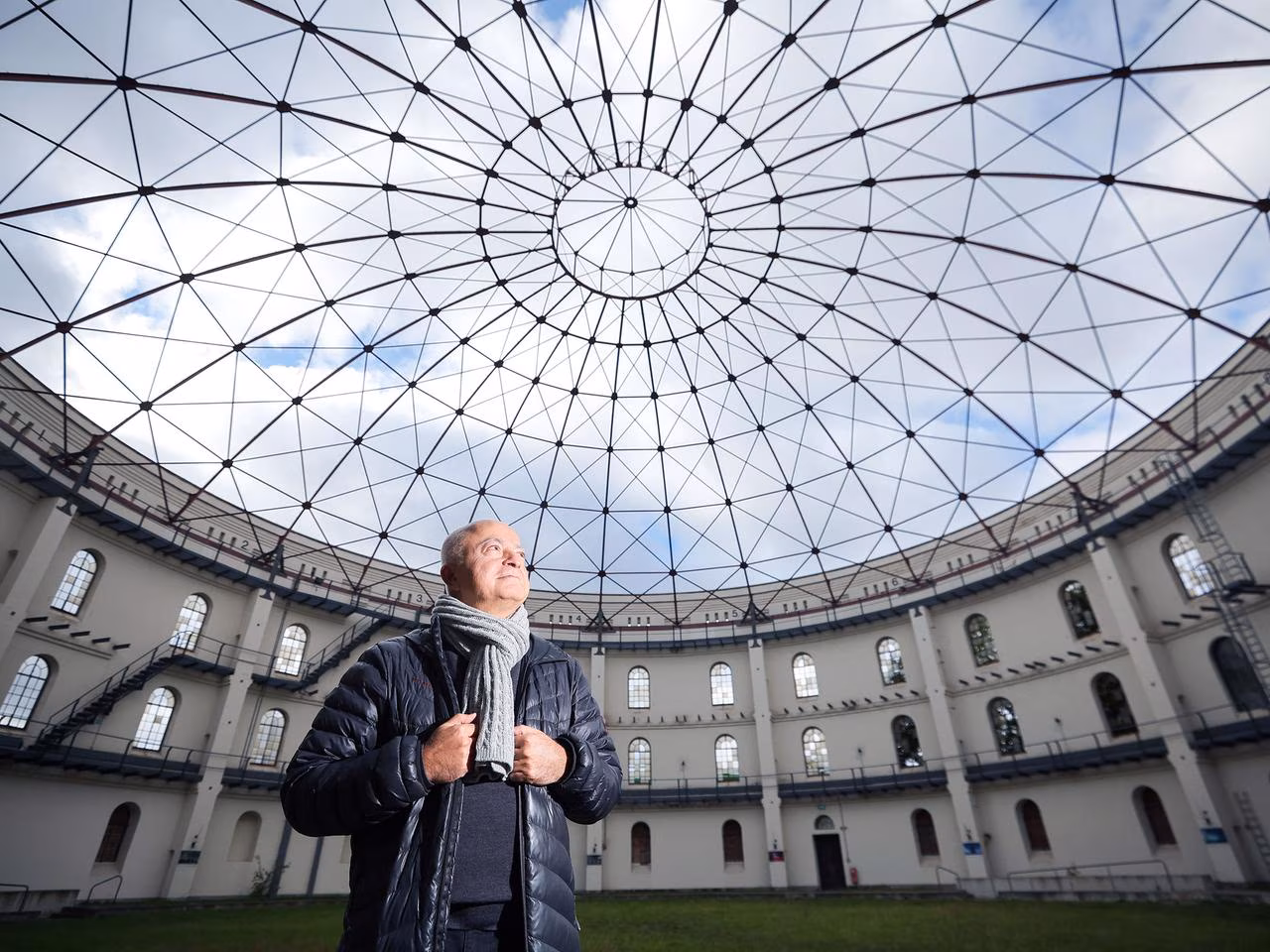 Artist Yadegar Asisi stands inside a panoramic building beneath a wide-span, delicate roof structure.